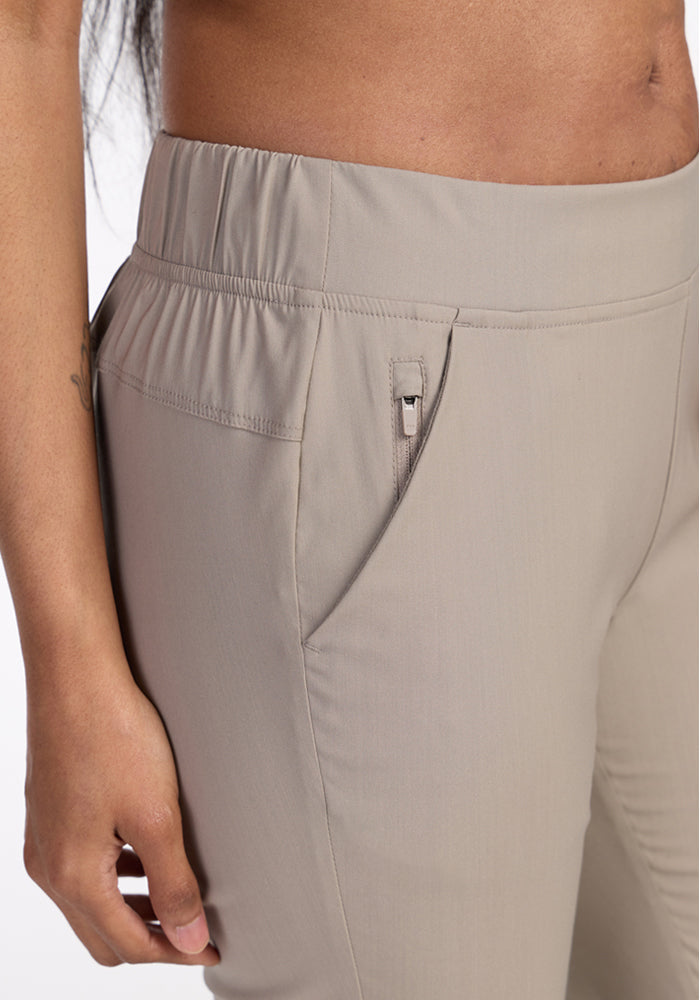Close-up of a person wearing Woolx Nicole Ankle Pants in Vintage Khaki, featuring a lightweight, breathable fabric, elastic waistband, and a small zippered pocket on the right side; their left arm rests by their side.