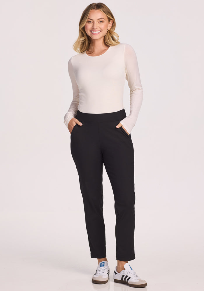 A woman smiles with her hands in the pockets of her Woolx Nicole Petite Ankle Pant in black, paired with a long-sleeve white top and white sneakers with black accents, standing against a plain light background.