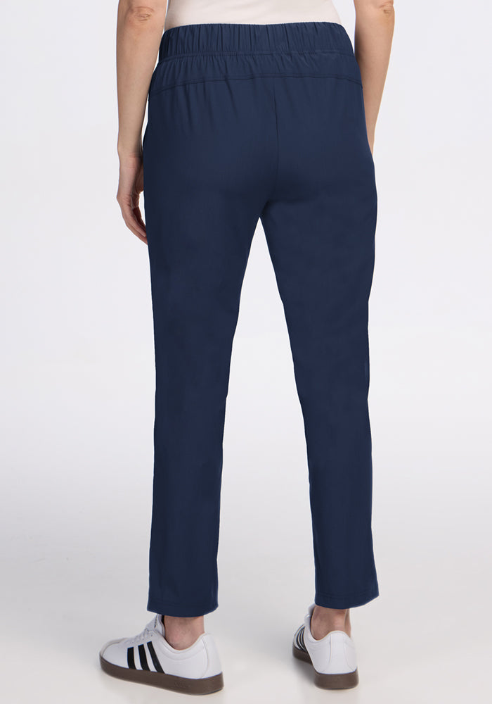 A person is shown from the waist down, back view, wearing Woolx Nicole Petite Ankle Pant in Deep Navy with white sneakers featuring black stripes, standing against a white background.