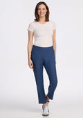 A woman with straight, shoulder-length brown hair poses casually against a white background, wearing a white tee, Woolx Nicole Petite Ankle Pant in Vintage Indigo, and white sneakers. She smiles warmly, showcasing a relaxed style.