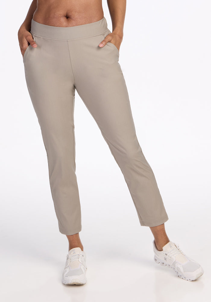 A person wears the Woolx Nicole Petite Ankle Pant in Vintage Khaki, styled with white sneakers and hands in pockets, standing against a plain white background.