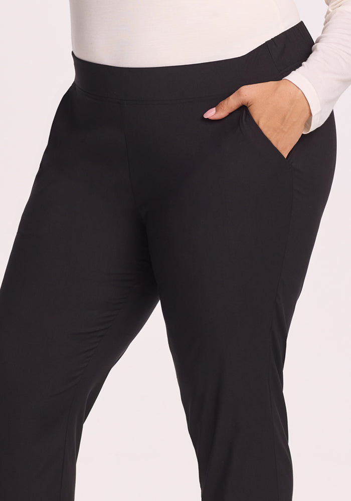 A person wearing the Woolx Nicole Tall Ankle Pant in black and a long-sleeved white top stands with one hand in their pocket against a plain light background.