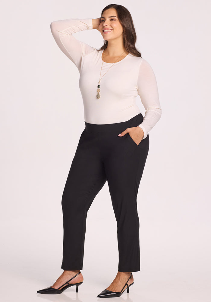A woman stands smiling with one hand on her hip, wearing a white long-sleeve top, black Woolx Nicole Tall Ankle Pant, black heels, and a long pendant necklace against a plain background.
