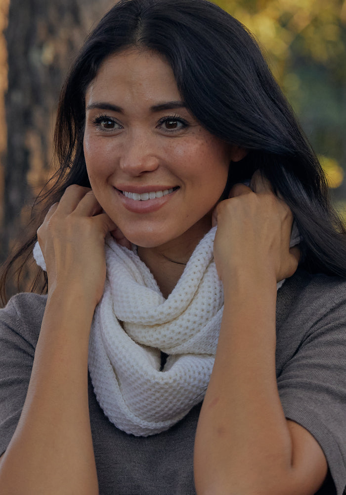 A woman with long dark hair smiles while wearing the Woolx Noelle Infinity Scarf in black around her neck. She pairs it with a gray top, and blurred trees with sunlight are visible in the background.