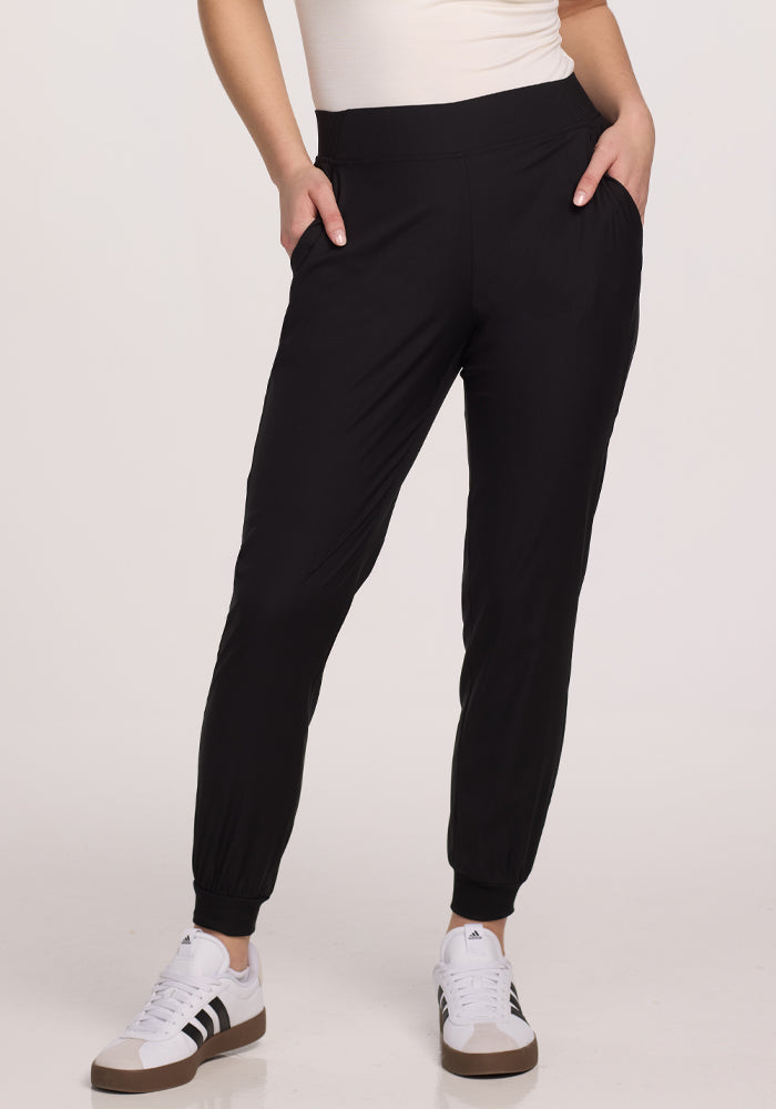 A person dressed in Woolx's black Palmer Joggers made from Merino fabric and white sneakers with black stripes stands against a plain backdrop. Their hands rest in the deep pockets, and they sport a light-colored top. | Ariana is 5'11", wearing a size S