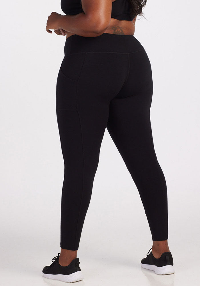 A person is standing with their back to the camera wearing Woolx's Piper Pocket Leggings in black and sneakers. The leggings feature moisture-wicking properties, making them an ideal cold-weather baselayer against the plain white background.
