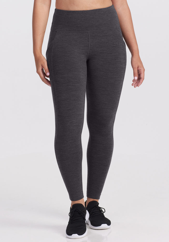 A person wears Woolx Piper Pocket Petite leggings in Cinder Melange, standing against a white background. These dark gray, high-waisted cold-weather leggings feature side seams and a smooth texture, perfect for warmth in chilly weather.