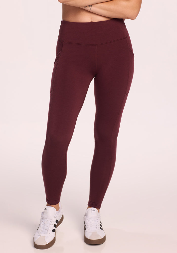 A person stands against a plain light background wearing Woolx Piper Pocket Petite leggings in Toasted Garnet with side pockets and white sneakers. The photo is cropped below the shoulders.