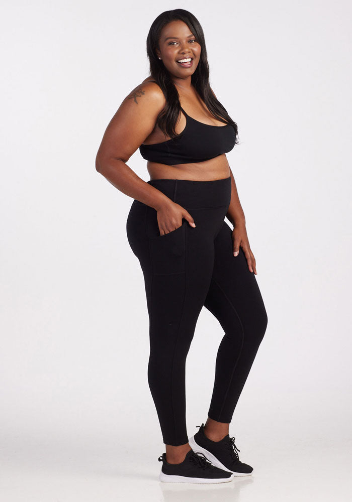 A woman smiling and posing against a white background is wearing the Piper Pocket Tall - Black sports bra from Woolx. Accompanying her look are high-waisted leggings with side pockets and black sneakers. Her long hair accentuates the tattoo on her upper arm.