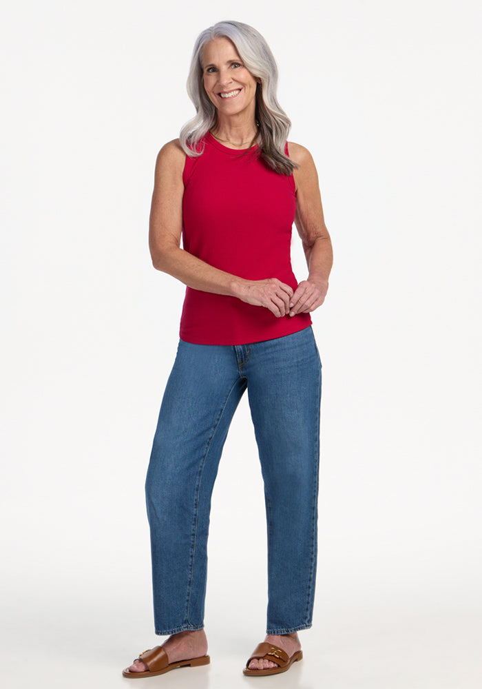 Smiling older woman wears Woolx Raegan Ribbed Tank Top in Poppy Spark, blue jeans, and brown sandals, standing on white.