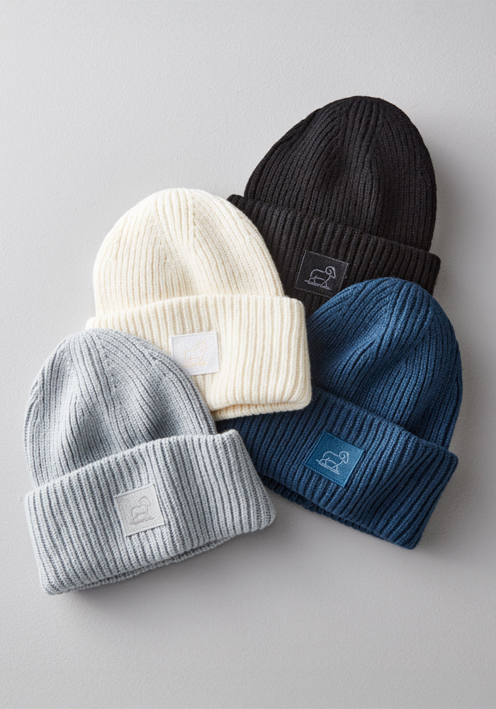 Four Woolx Ria Chunky Beanies in black, cream, light gray, and dark blue are displayed on a light surface. Each ribbed merino wool beanie features a folded brim with a matching rectangular logo patch.