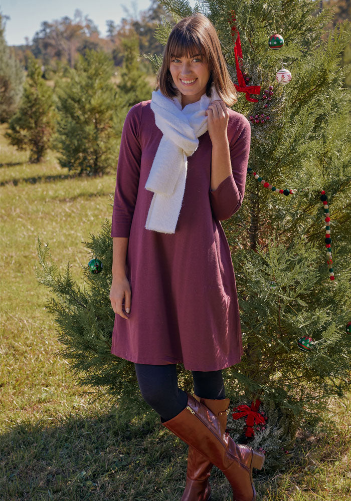 A woman in the Woolx Rissa A Line Dress - Cinder Melange, black leggings, brown boots, and a white scarf smiles beside an outdoor Christmas tree—a stylish fall and winter look with trees and grass in the background.