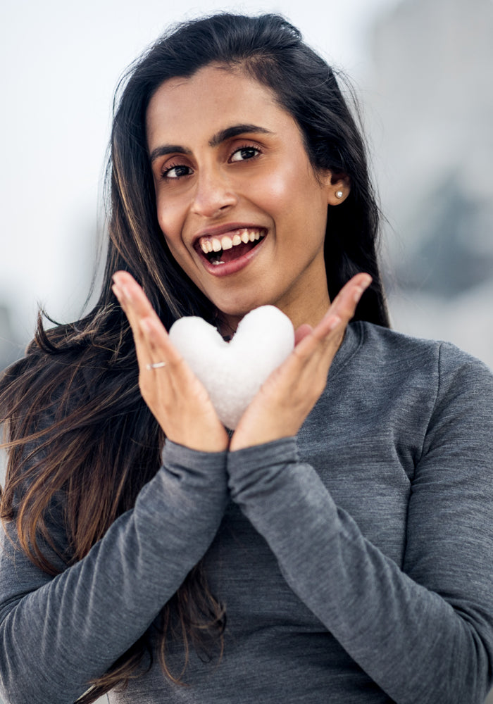 A smiling woman with long dark hair wears the Woolx Sadie Baselayer Top in Black as she holds a heart-shaped snowball in her cupped hands. The softly blurred background highlights her cozy top and joyful expression.
