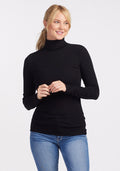 A person with blonde hair stands against a light background, wearing the Sage Ribbed Turtleneck in black by Woolx and blue jeans. The moisture-wicking fabric of the turtleneck ensures comfort. They have a relaxed expression and their hands are gently clasped in front.