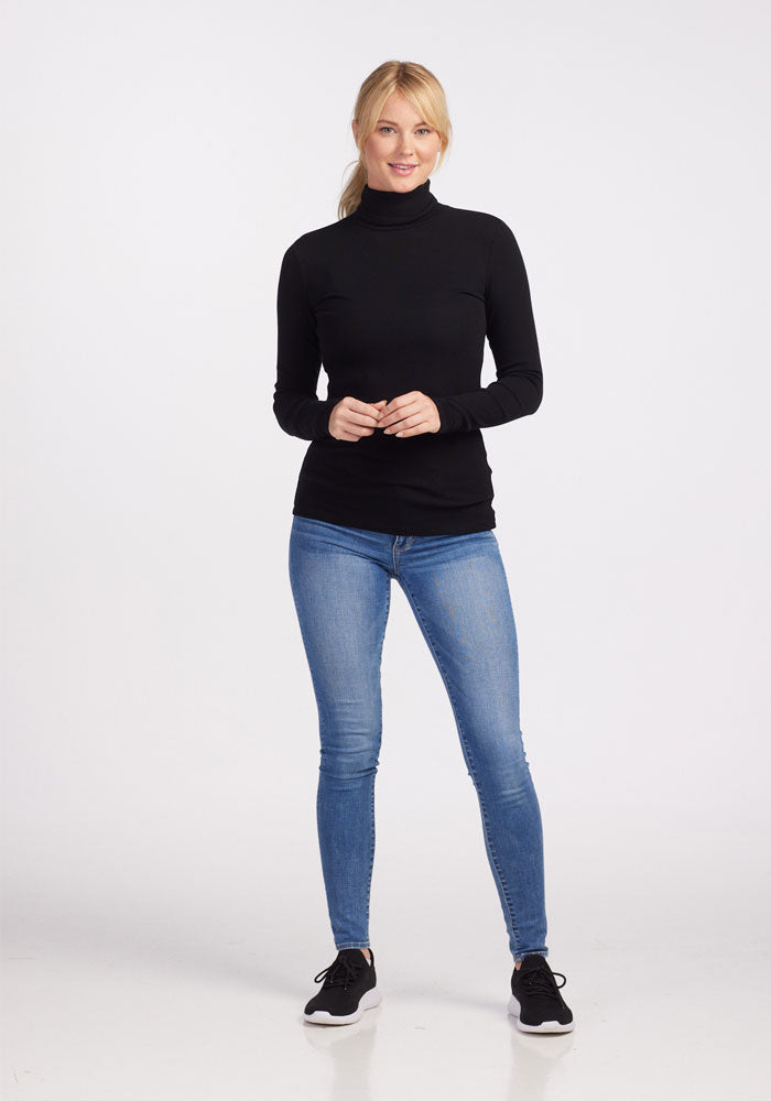 A person stands against a plain white background wearing a black Sage Ribbed Turtleneck from Woolx, blue jeans, and black sneakers. They have blonde hair and are smiling with hands clasped in front. This moisture-wicking attire combines comfort and style seamlessly.