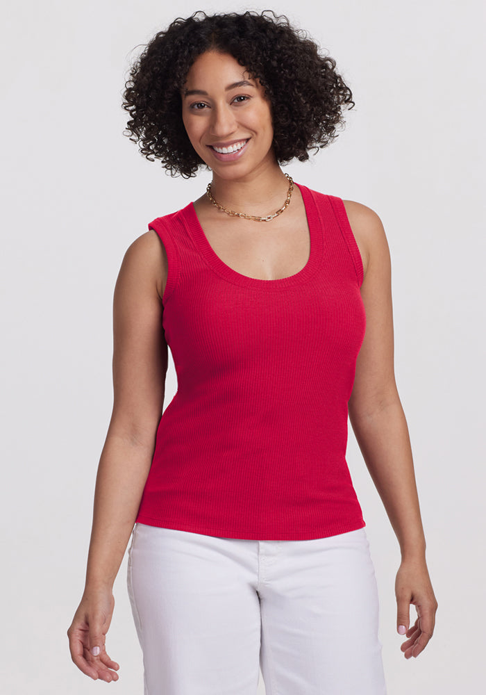 Smiling woman in Woolx Sloane Ribbed Tank in Poppy Spark and white pants stands by a plain white background.
