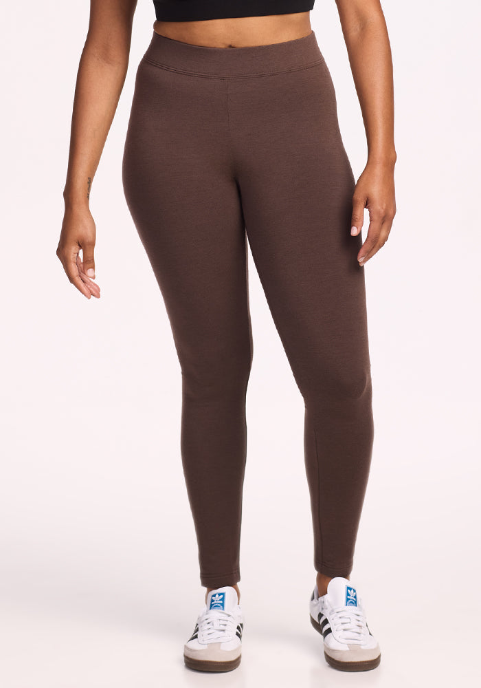 A person stands against a plain light background, wearing Woolx's Stella Petite merino wool leggings in Bracken and white sneakers with blue accents. The photo is cropped from the waist down.