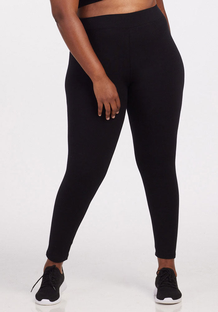 A person wearing Woolx's Stella Tall leggings in black stands against a plain white background. They are visible from the waist down, with their right hand resting on their thigh, highlighting the sleek design of these cozy baselayer leggings paired with black sneakers.