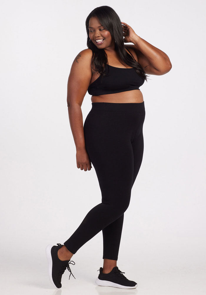 A person is smiling, wearing a black sports bra, tall-sized Stella Tall - Black leggings by Woolx made from DuraLite fabric, and black sneakers. They are standing in a relaxed pose against a plain white background.