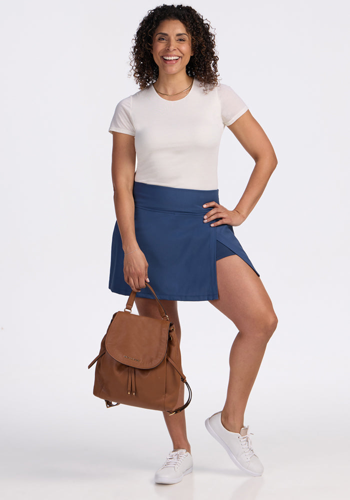 A smiling woman with curly hair holds a brown backpack. She wears a white tee, Woolx Topanga Skort in Vintage Indigo with a front slit, and white sneakers against a plain white background.