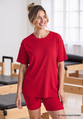 Woman in the Woolx Vera Ribbed Tunic in Poppy Spark smiles in a bright room with Pilates equipment.
