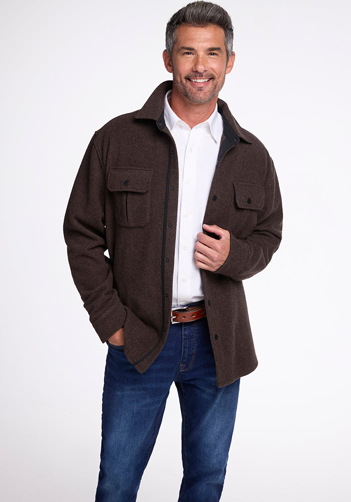 A man with gray hair smiles while standing in a relaxed pose, wearing the Woolx Wilder Shirt Jac in Maple Melange over a white button-up shirt crafted from Merino wool. He pairs this ensemble with blue jeans and a brown belt, embracing an ultra-functional style against a plain white background.