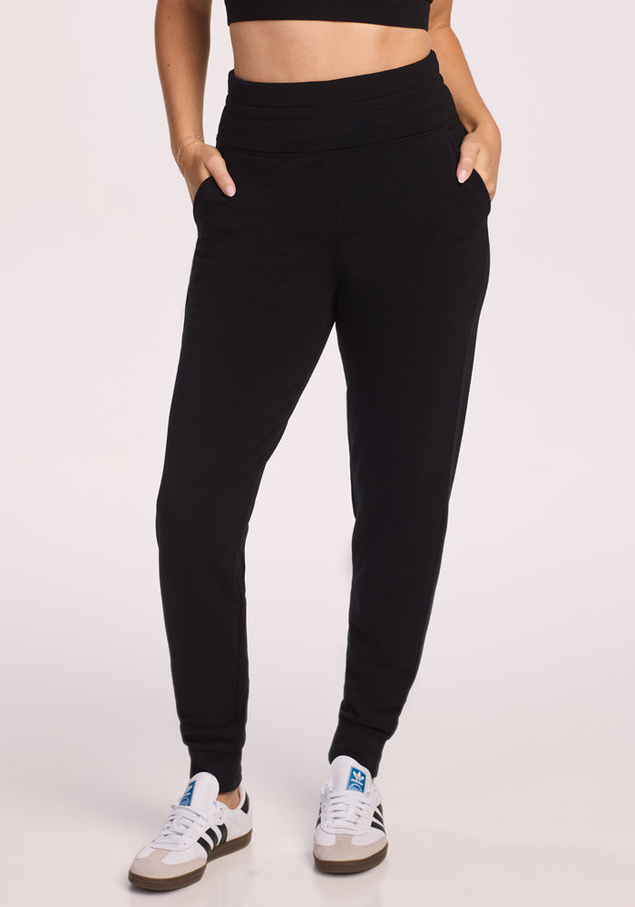 A person stands with hands in pockets against a light background, wearing Woolx Willa Joggers Petite in black, paired with a black crop top and white sneakers with black stripes.