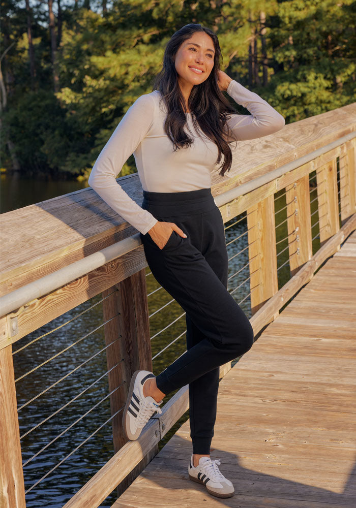 A woman with long dark hair, in a light long-sleeve top, white sneakers, and Woolx Willa Joggers Petite, smiles and leans against a wooden bridge railing over a lake with trees in the background.