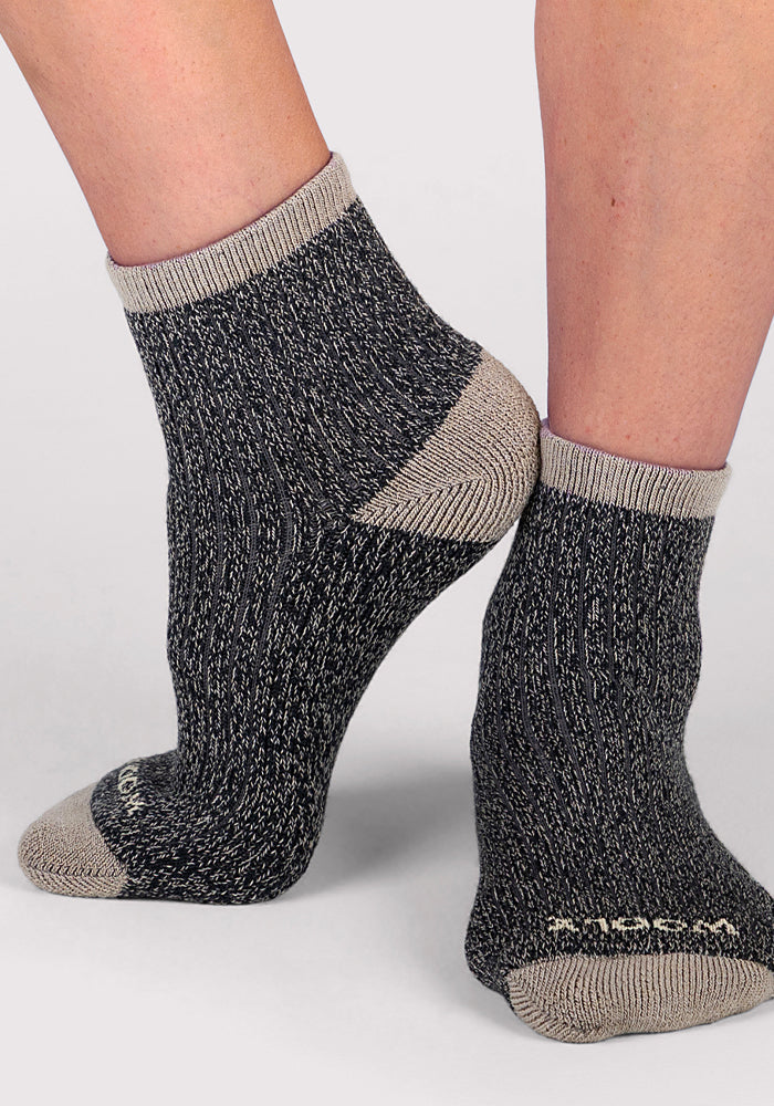 A close-up of a person wearing Woolx Womens Cozy Mini Crew Sock in black, featuring ribbed Merino wool with contrasting heels, toes, and cuffs, standing on a light background.