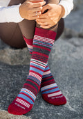 A person sits on a rock wearing Woolx Womens Everyday Crew Socks in Rhubarb Stripe—colorful, moisture-wicking Merino wool socks featuring red, blue, gray, and white stripes with "Woolx" branding on the toes. Their legs are crossed and hands rest on their knee.