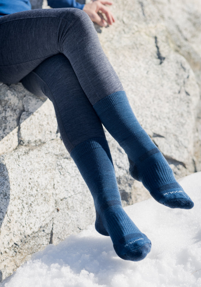 A person sits cross-legged on a rock with snow around, wearing dark blue and grey leggings and Woolx Womens Harbor Crew Socks in blue.