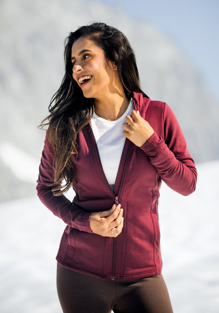 A woman with long dark hair wears a maroon Woolx Zoey Zip Up Hoodie over a white shirt, standing outdoors in a snowy mountain setting, smiling and looking to the side.